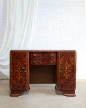 Load image into Gallery viewer, A vintage Art Deco writing desk with rich reddish-brown burl wood grain, featuring two side cabinets, a central drawer, and a dark open center compartment, set against a white brick wall and draped white fabric.
