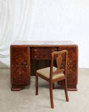 Load image into Gallery viewer, An Art Deco style writing desk with a burl wood veneer finish and a marble top, accompanied by a matching wooden chair with a beige upholstered seat, set against a white brick wall and draped white fabric.
