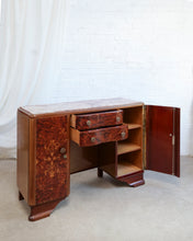 Load image into Gallery viewer, An Art Deco style wooden writing desk or credenza with a burl wood cabinet door, two drawers, open shelving, and an open side door, set against a white brick wall and white curtain backdrop.
