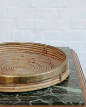 Load image into Gallery viewer, A round woven rattan tray with a polished brass rim, shown resting on a dark green marble slab against a white brick wall background.
