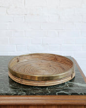 Load image into Gallery viewer, A round woven rattan tray with a polished brass rim, displayed on a dark green marble-topped table against a white brick wall background. Italian Rattan Tray In The Style of Gabriella Crespi
