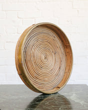 Load image into Gallery viewer, A round woven bamboo tray with a spiral pattern, standing on a dark marble surface against a white brick wall background.
