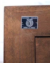 Load image into Gallery viewer, Close-up of a wooden cabinet corner with a small metal plaque reading 'NAVARRO ARGUDO MADE IN SPAIN' and a central 'NDA' emblem.
