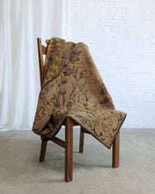 Load image into Gallery viewer, A vintage-style tapestry throw blanket featuring a dog and floral scrollwork pattern in earthy brown and beige tones, draped over a wooden chair in a bright room with a white brick wall and white curtain.
