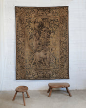 Load image into Gallery viewer, A large vintage tapestry featuring a hunting dog and a stag in a forest scene, displayed on a white brick wall above two small wooden stools.
