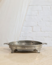 Load image into Gallery viewer, A small round metal tray with side handles and three short feet, shown on a light stone surface against a white brick wall background.
