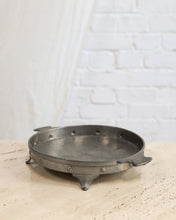 Load image into Gallery viewer, A round, dark gray cast iron trivet or burner base with three legs and rivet details, resting on a light beige stone surface against a white brick wall background.
